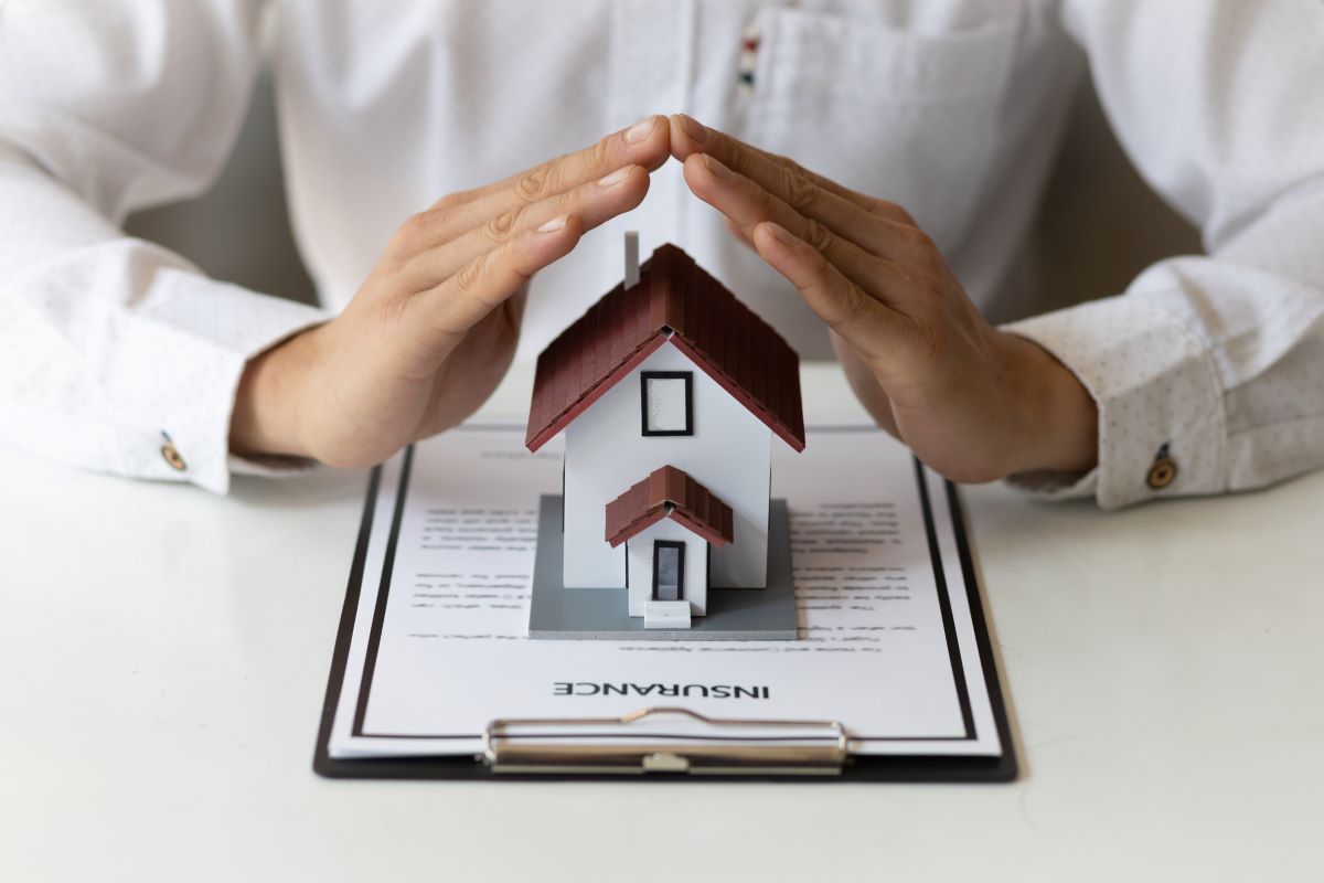 A person's hands are arched over a small model of a home that is placed on top of an insurance contract.